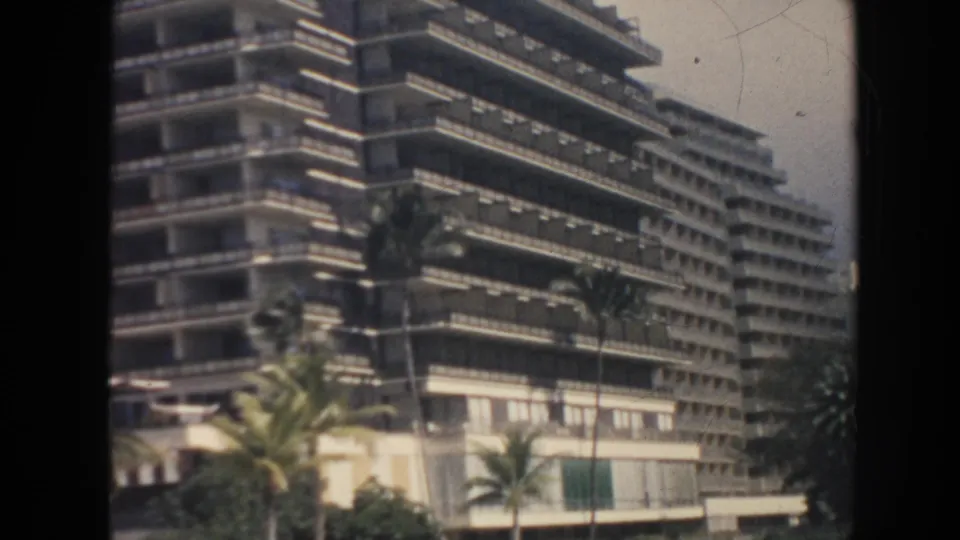 1961 Hawaii home-movie frame of a mid-century resort-style building with stacked balconies and palm trees in the foreground.