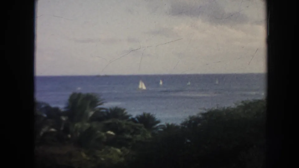 1961 Hawaii coastal frame with cloudy sky, blue ocean, small sailboats, and dark tropical foliage in front.