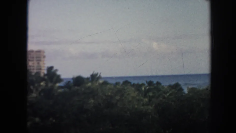 1961 Hawaii 8mm frame showing dense tropical greenery, palm tops, blue ocean, cloudy sky, and a distant shoreline building at left.
