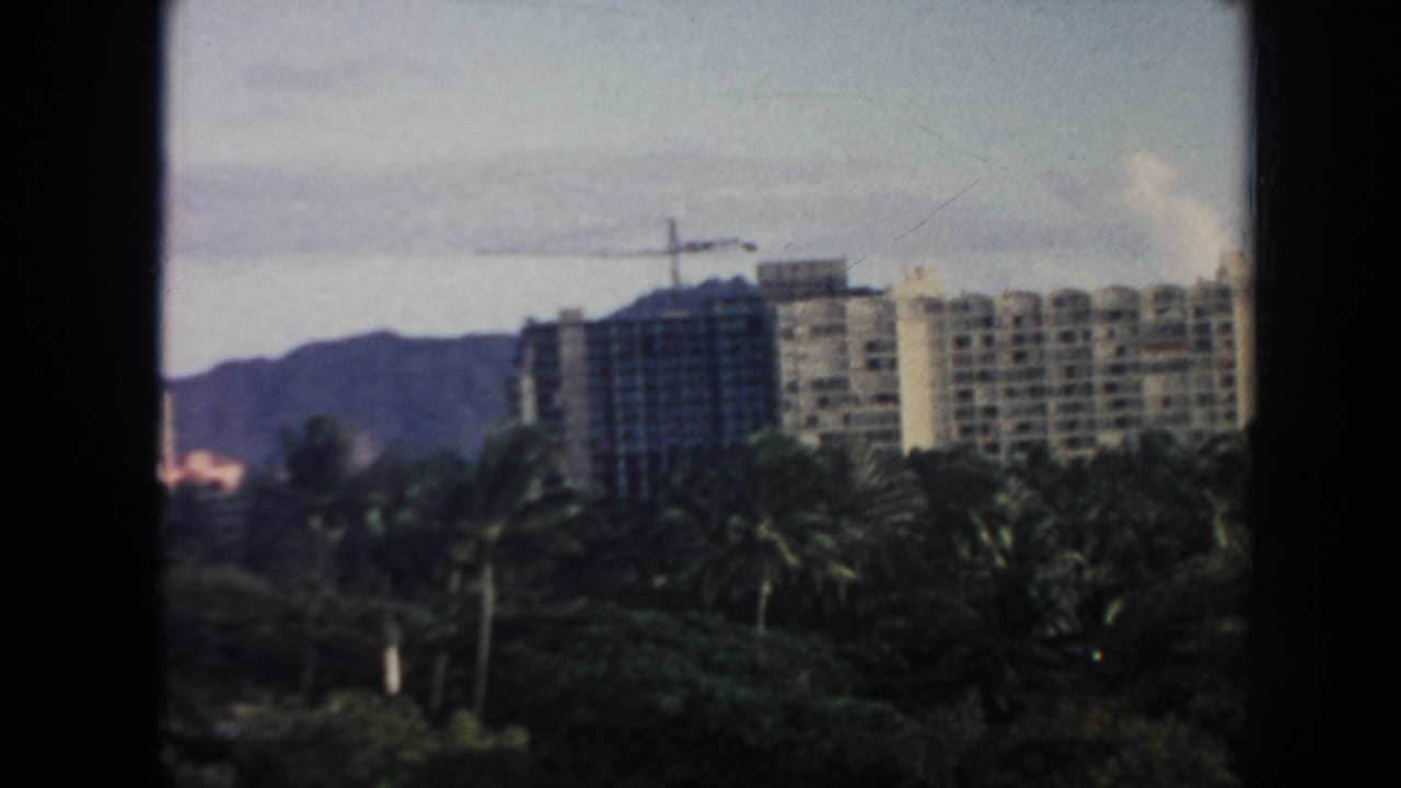 High-rise buildings, a crane, palm trees, and mountains seen through a dark window frame.