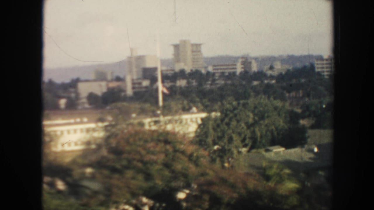 1961 Hawaii elevated view with white buildings, trees, flagpoles, and distant high-rises under hazy daylight.