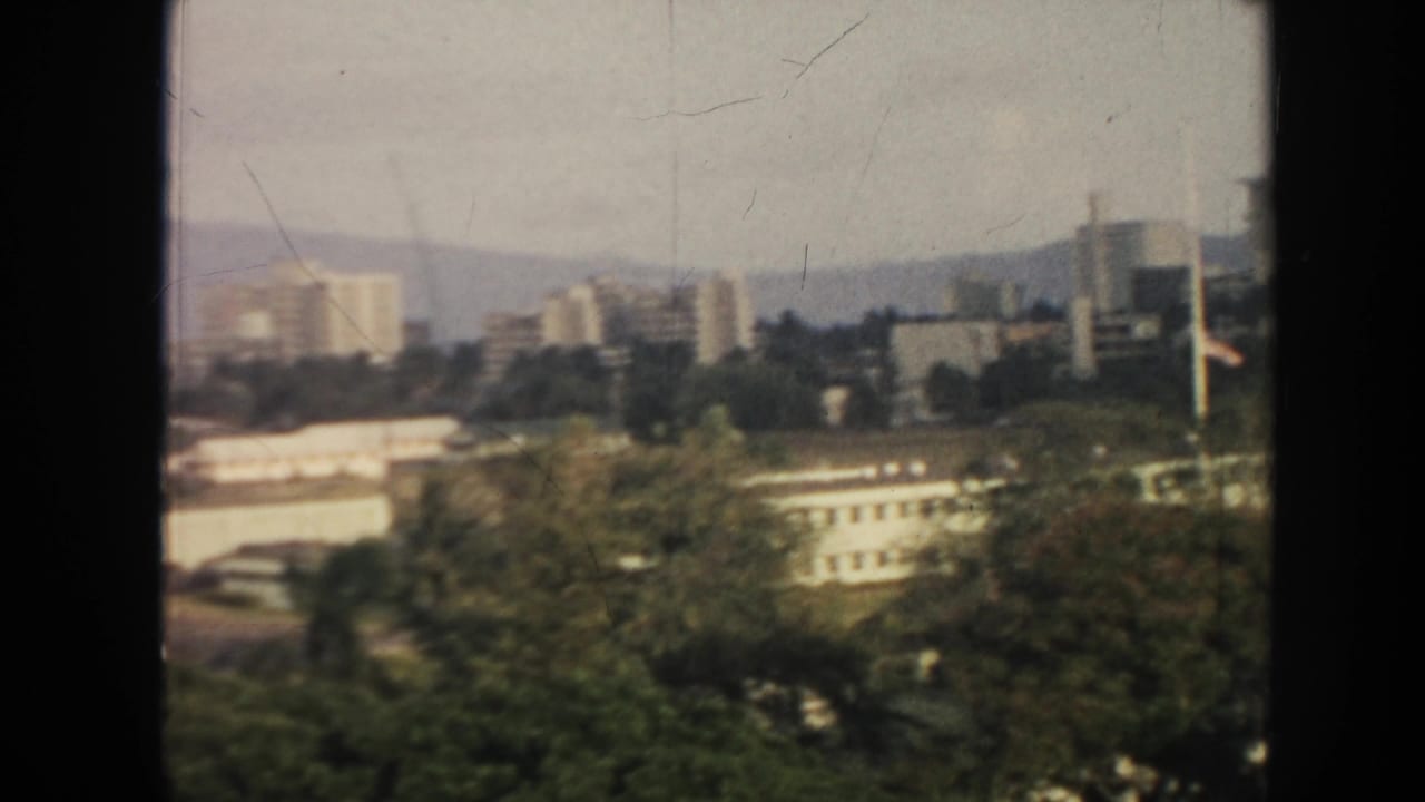 1961 Hawaii home-movie frame showing a hazy cityscape of white buildings, trees, flagpoles, and distant mountains.