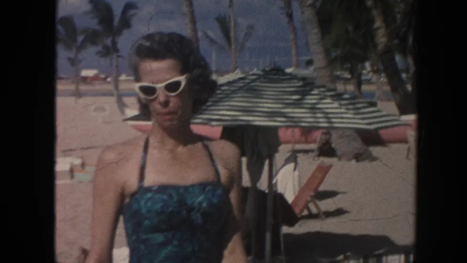 1961 Hawaii beach frame showing an adult woman posing near a striped umbrella, with palm trees in the background.