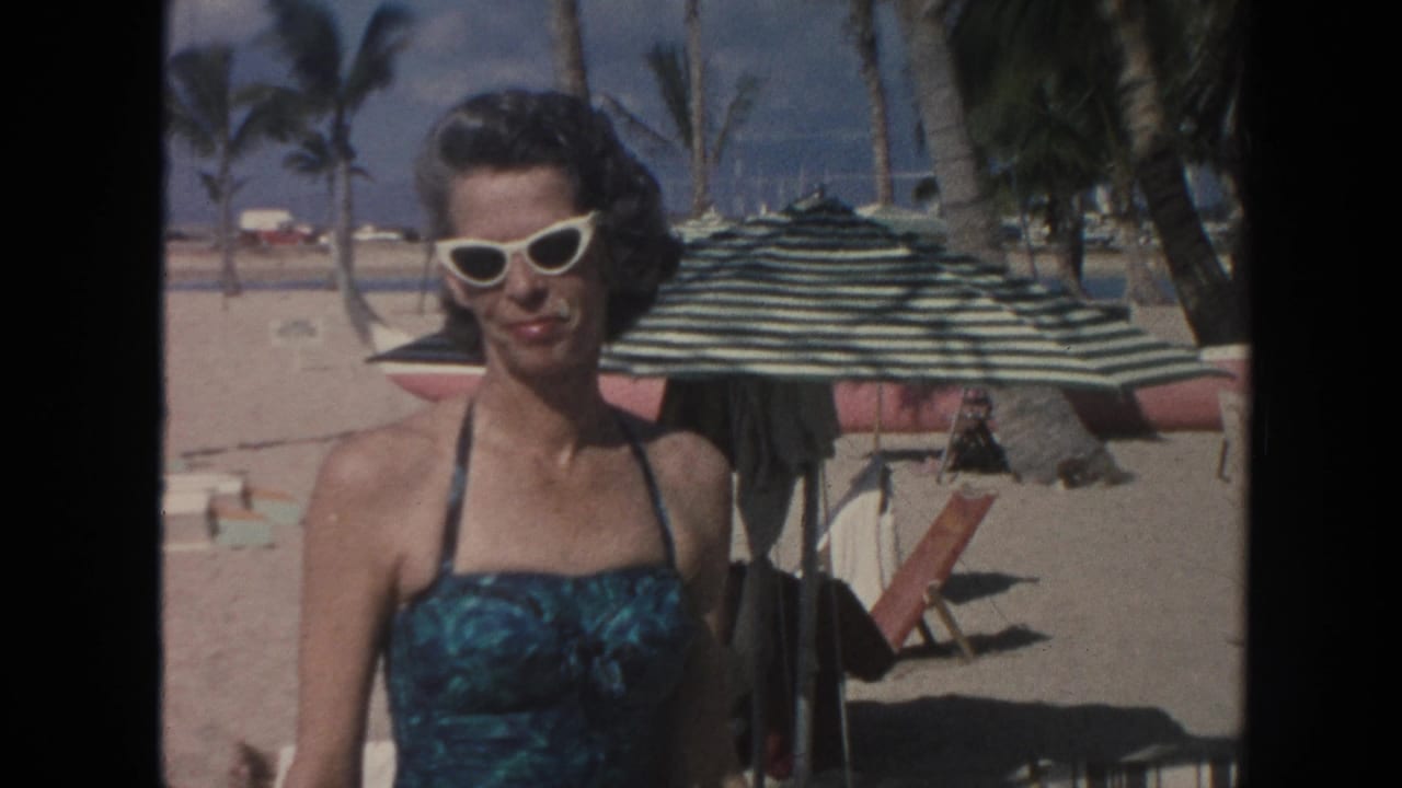In 1961 Hawaii home-movie footage, an adult woman in a teal swimsuit and white cat-eye sunglasses faces the camera near a beach umbrella.