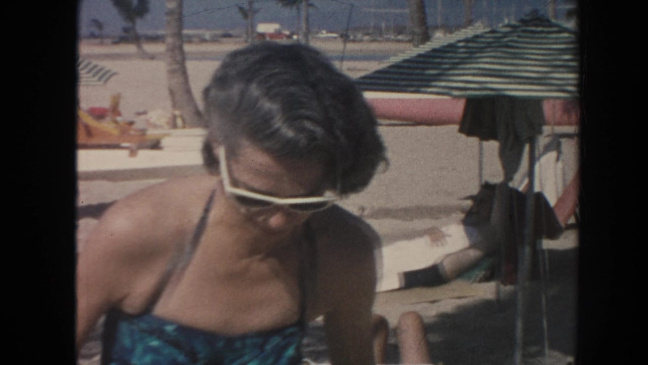 The woman in a teal swimsuit and white sunglasses looks down as she moves beside a striped beach umbrella on a Hawaii beach.