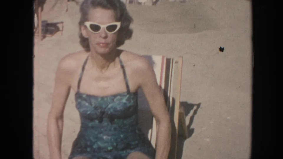An adult woman in white cat-eye sunglasses and a teal swimsuit sits on a striped lounge chair in 1961 Hawaii beach footage.