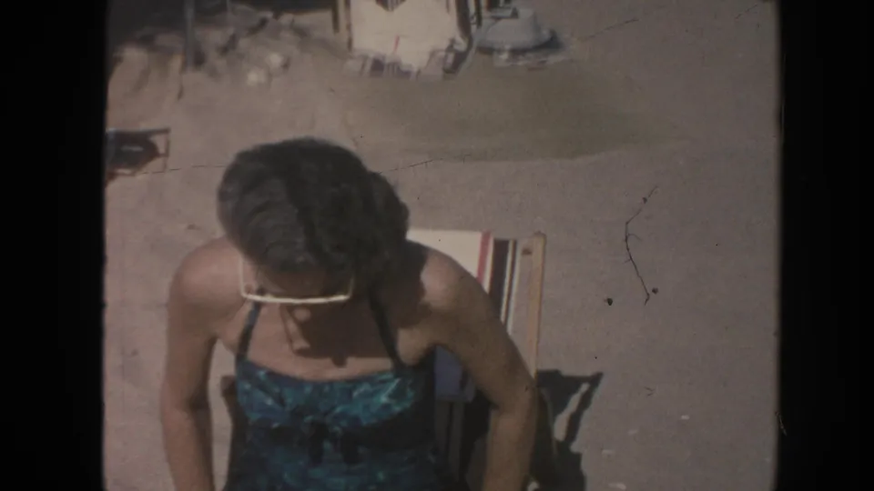 The woman leans forward and looks down while seated on a striped lounge chair during a 1961 Hawaii beach home movie.