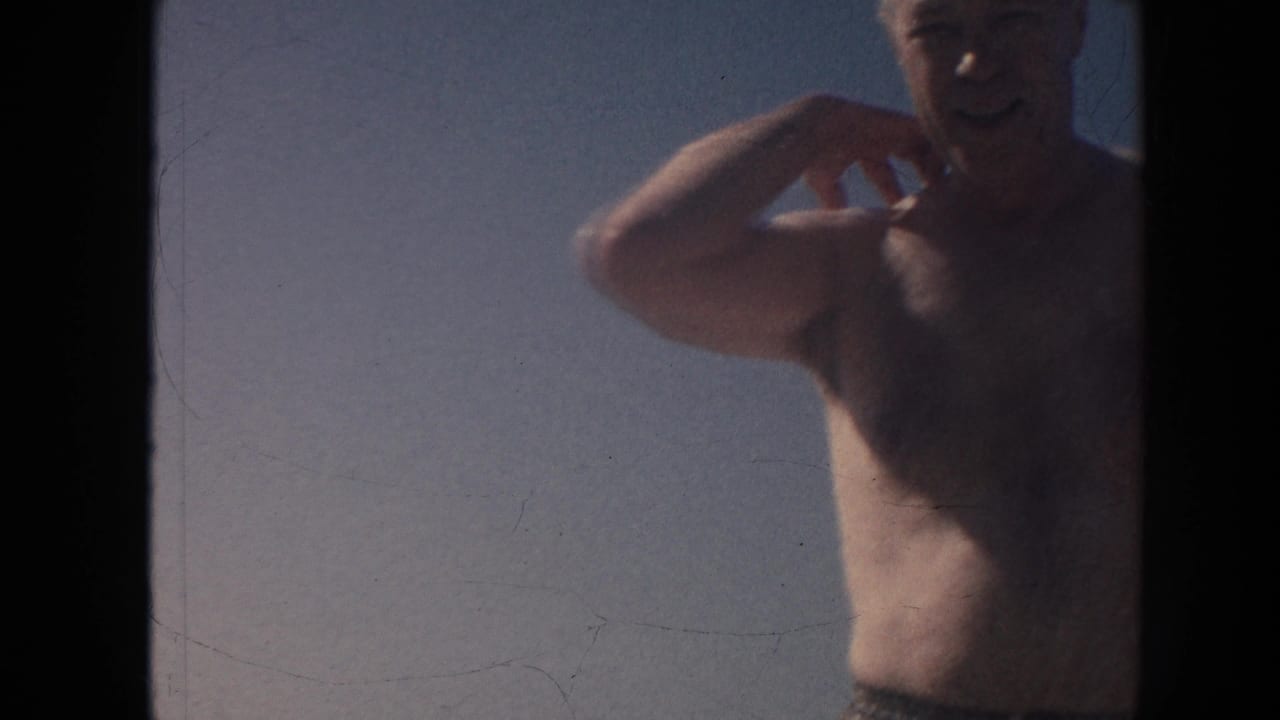 1961 Hawaii home-movie frame of a shirtless older man smiling and gesturing near the beach under a blue sky.