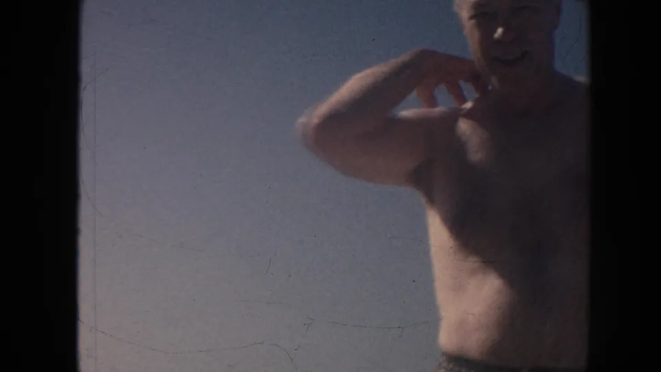 1961 Hawaii home-movie frame of a shirtless older man smiling and gesturing near the beach under a blue sky.