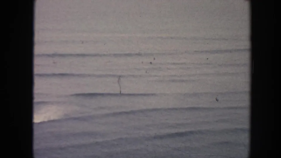 1961 Hawaii home-movie frame of soft ocean waves and pale horizon, marked by visible 8mm grain and scratches.