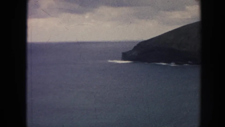 1961 8mm Hawaii home-movie frame showing a dark rocky headland or mountain slope beside open blue ocean with small surf marks under cloud cover.