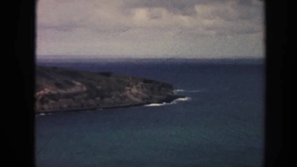 1961 8mm Hawaii coastal frame showing a low rocky peninsula bordered by blue-green ocean, white surf at the shoreline, and a cloudy horizon.