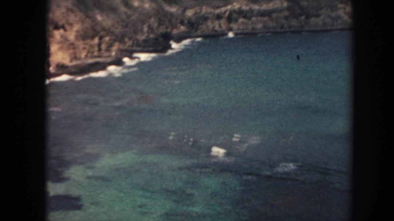 1961 Hawaii 8mm frame showing turquoise coastal water, dark reef or rock patches, and surf along a rugged shore.