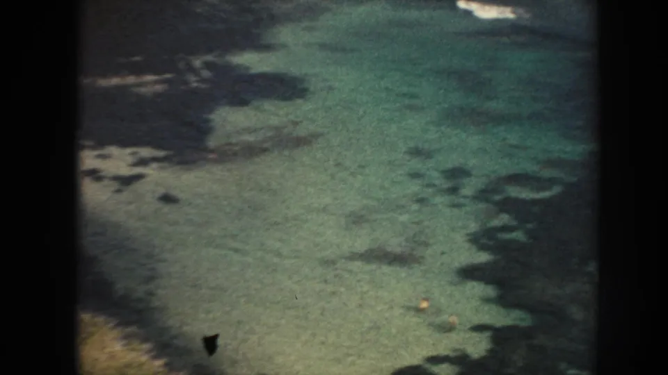 1961 Hawaii 8mm frame looking down into clear turquoise shallows with dark rock or reef forms near the beach edge.