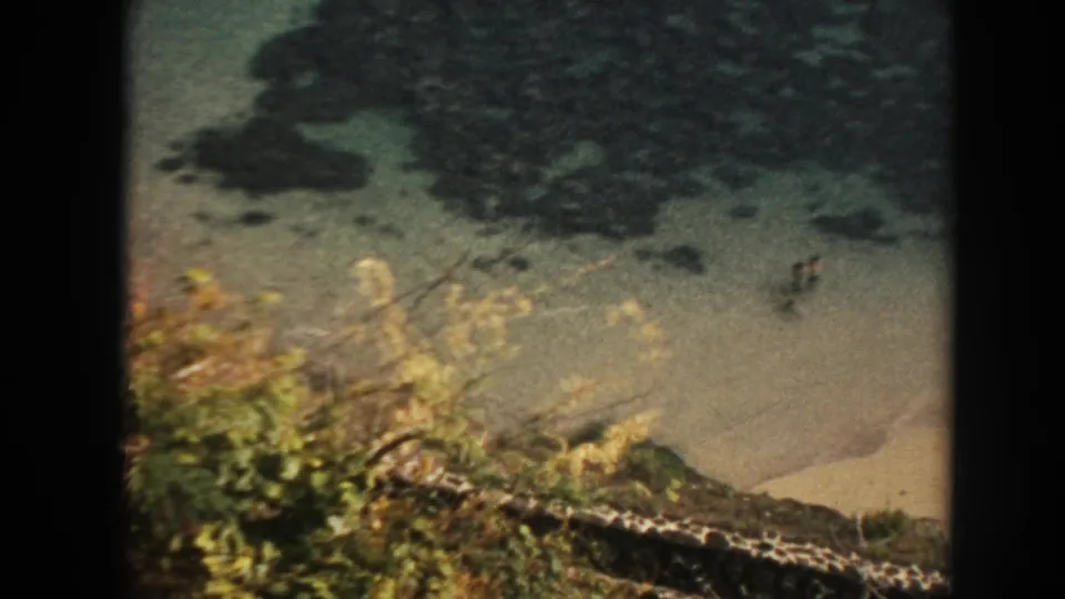 1961 Hawaii 8mm frame of a rocky beach, green foreground foliage, clear water, and two distant people wading in the surf.