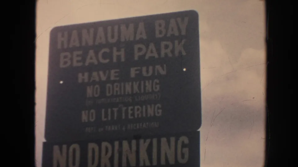 Close 1961 Hawaii 8mm frame of the Hanauma Bay Beach Park sign, with no drinking and no littering rules readable in soft focus.