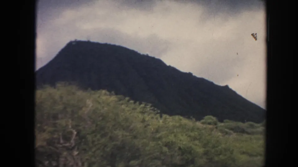1961 Hawaii home-movie frame of a volcanic hillside with green brush and overcast sky.