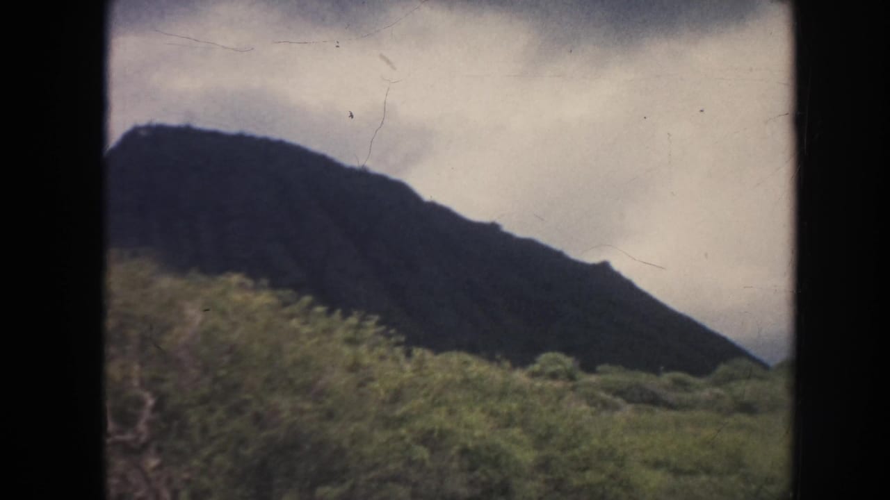 1961 Hawaii 8mm frame showing a dark mountain ridge rising behind dense green trees beneath a cloudy sky.