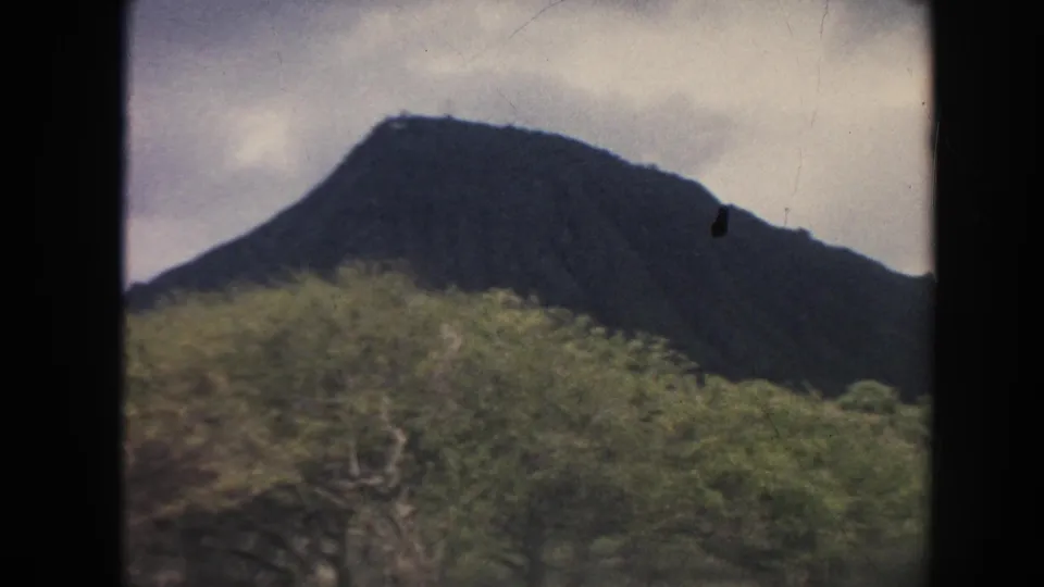 1961 Hawaii 8mm frame showing a sloping mountain silhouette beyond thick green foliage under clouded light.