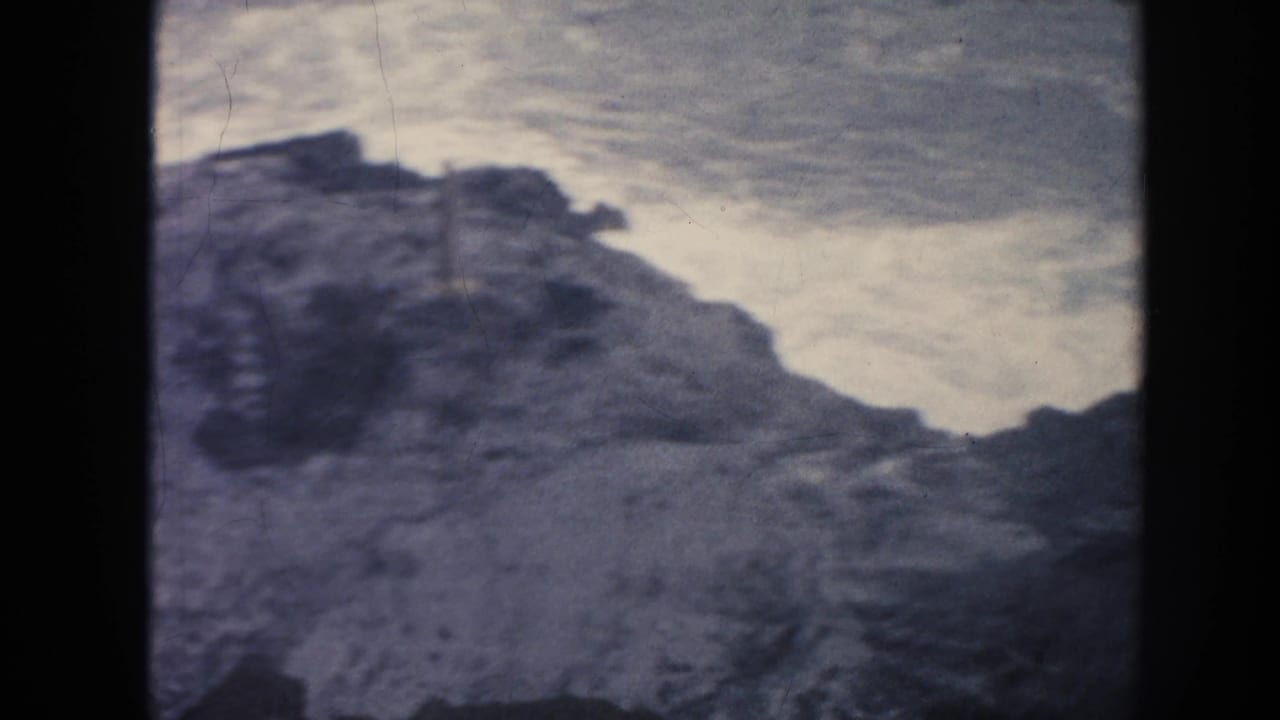 1961 Hawaii 8mm frame showing dark rock forms below pale, churning surf and overcast coastal light.