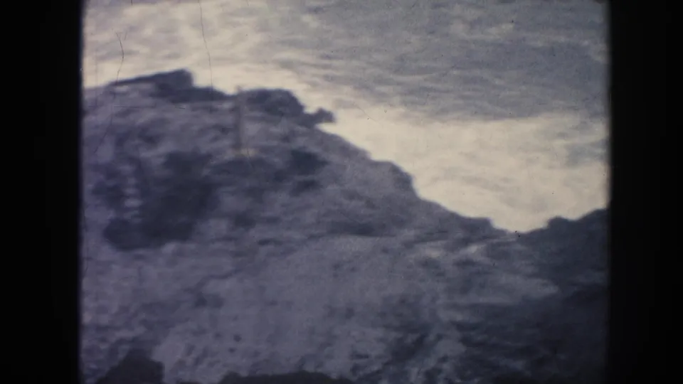 1961 Hawaii 8mm frame showing dark rock forms below pale, churning surf and overcast coastal light.