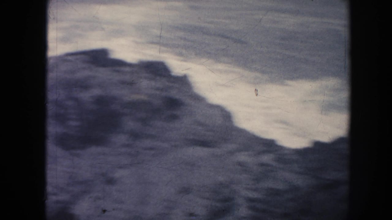 Rocky shoreline mass against pale moving water, with dark film border visible.