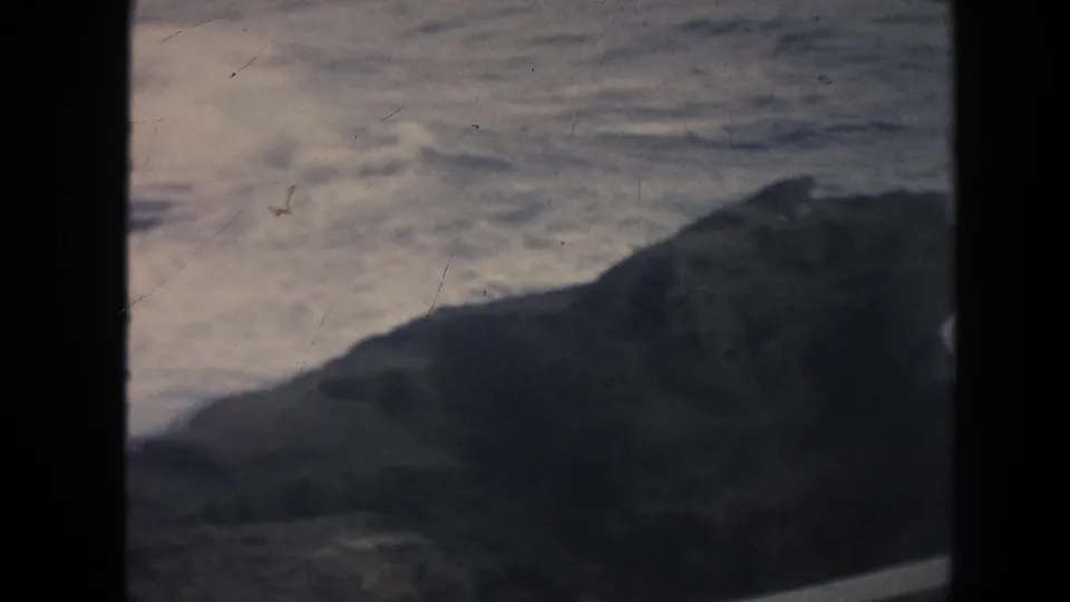 1961 Hawaii 8mm frame showing a dark sloping coastal rock beside pale rough water under muted light.