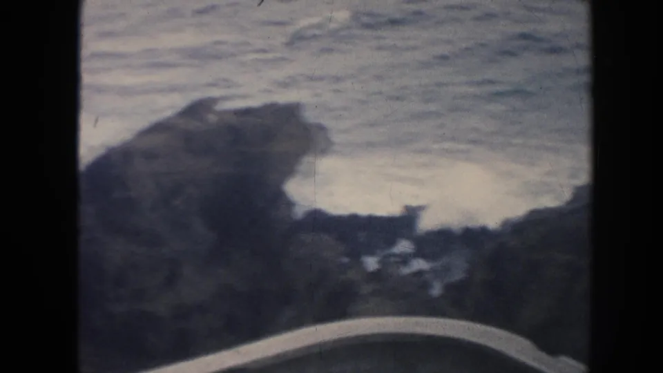 1961 Hawaii 8mm frame showing rough surf washing around dark jagged coastal rocks, with a pale curved foreground edge partly visible.