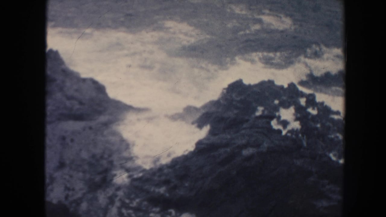 1961 Hawaii 8mm frame with white waves surging through dark coastal rocks under muted blue-gray film color.