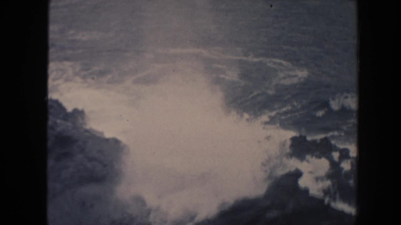 1961 Hawaii 8mm frame with bright ocean foam spreading around dark shoreline rocks, softened by grain, fading, and frame-edge shadow.