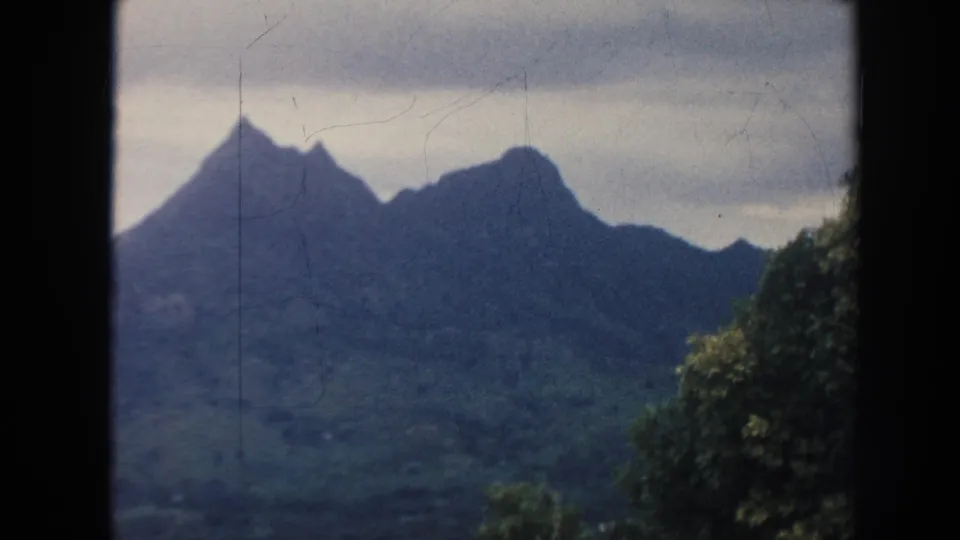 1961 Hawaii 8mm frame showing a green mountain range under low clouds, with jungle foliage along the right and lower edges of the view.