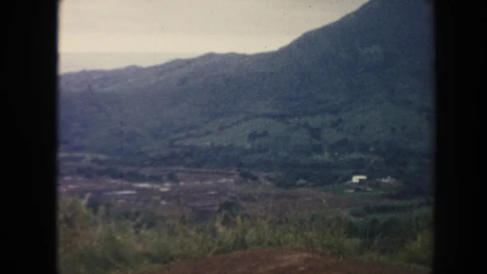 1961 Hawaii 8mm frame with rolling green hills, valley terrain, roadside vegetation, and a distant white building.