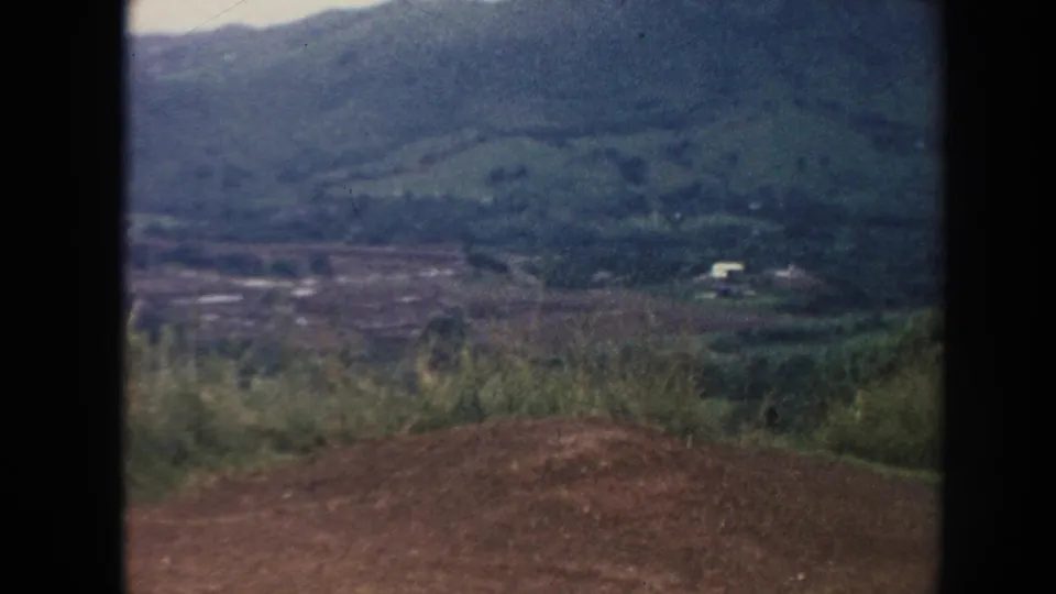 1961 Hawaii 8mm frame looking from a dirt roadside foreground across vegetation toward a green valley and distant building.