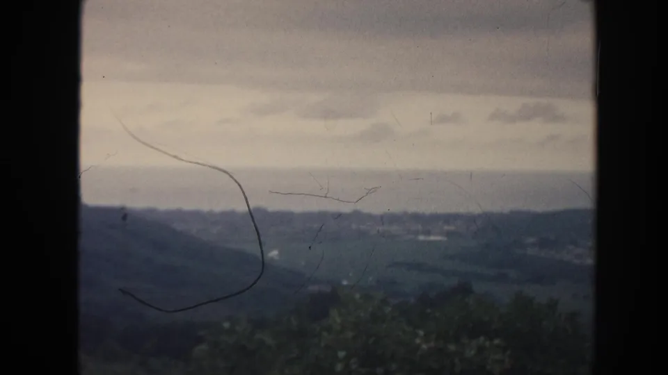1961 8mm Hawaii frame looking over dark green hills toward a pale horizon, with brush in front and scratches on the film.