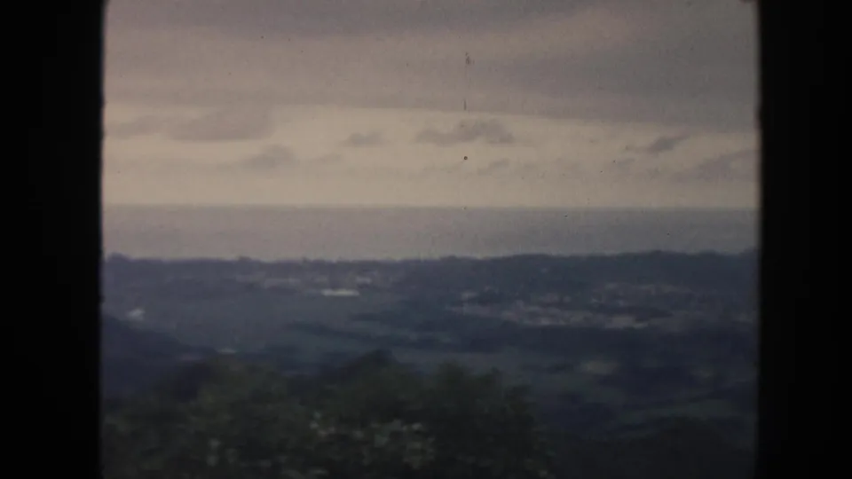 1961 8mm Hawaii frame showing a wide rural vista with green hills, distant buildings, foreground vegetation, and cloudy sky.