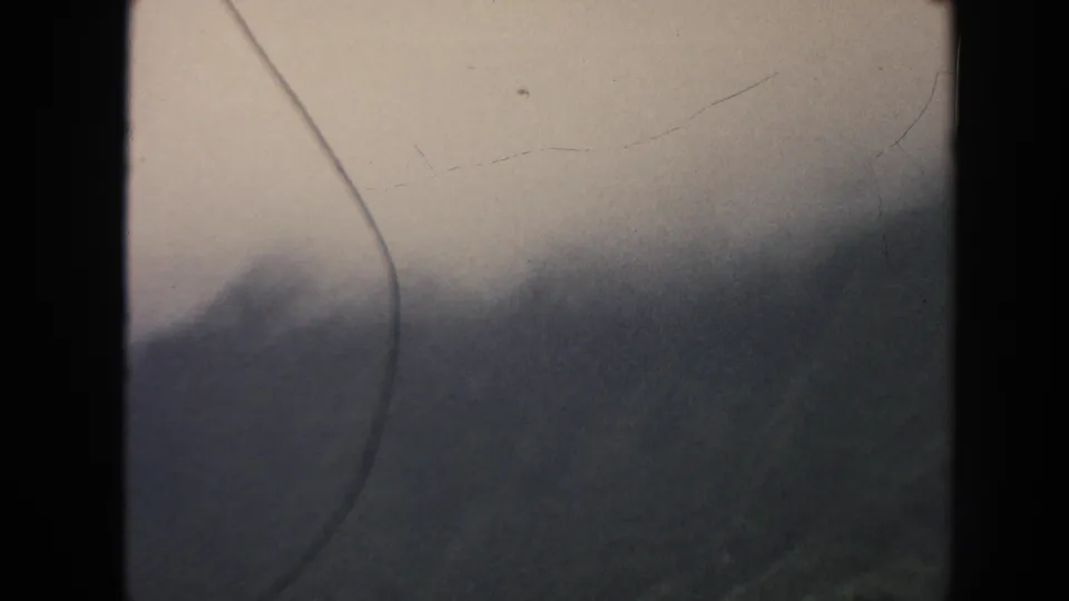 1961 Hawaii 8mm frame showing a mist-covered mountain slope, dim forest texture, pale sky, and aged film surface marks.