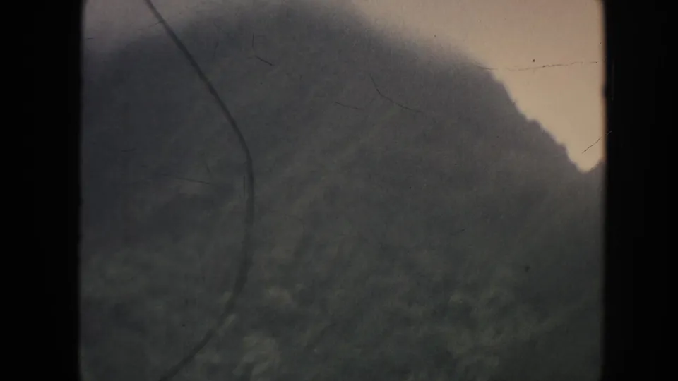 1961 Hawaii 8mm frame with a forested mountain side, cloud-softened ridge line, pale sky, and visible aged-film scratches.