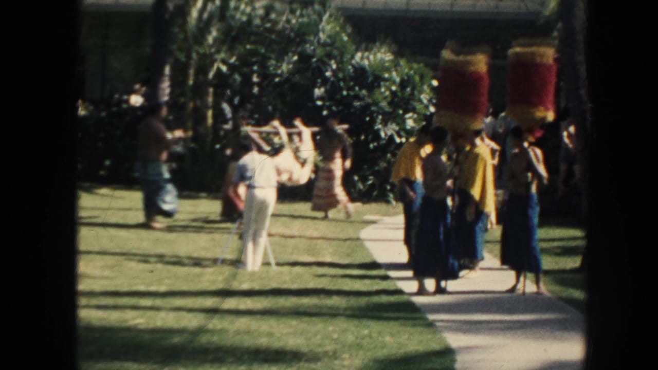Performers and tourists watch men carrying an animal on a pole across a lawn.
