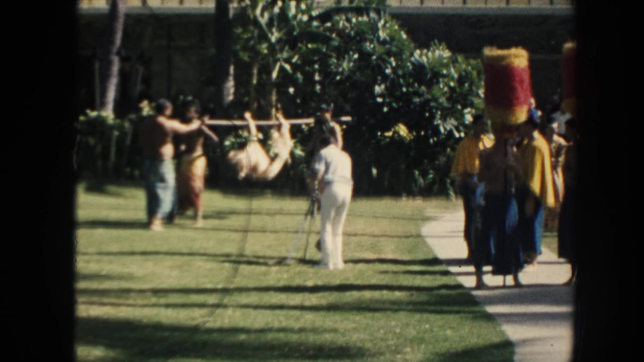 1961 Hawaii garden scene with visitors photographing men carrying a tied animal on a pole near performers in tall red-and-yellow headpieces.