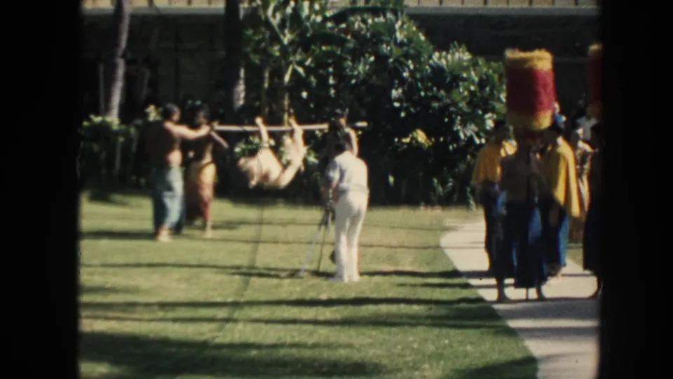 1961 Hawaii garden scene with visitors photographing men carrying a tied animal on a pole near performers in tall red-and-yellow headpieces.