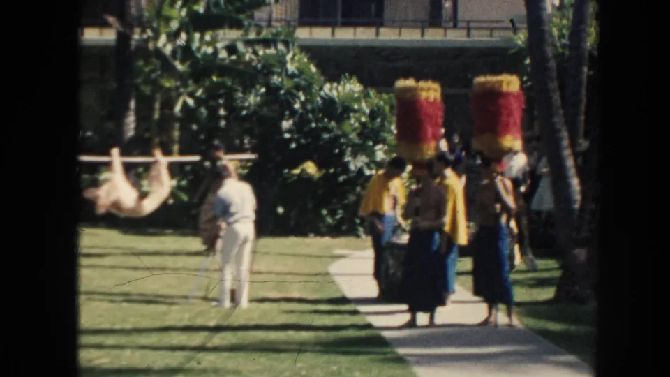 1961 Hawaii home-movie frame with costumed performers, visitors, and men carrying an animal on a pole through a tropical lawn setting.