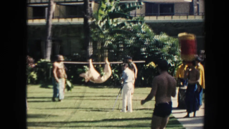 1961 Hawaii frame showing a shirtless foreground figure, tourists, performers, and a tied animal carried on a pole in a garden ceremony setting.
