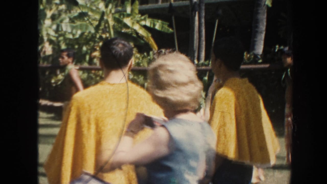 1961 Hawaii home-movie frame showing tourists from behind as men in yellow capes move near a pole in a lush outdoor setting.