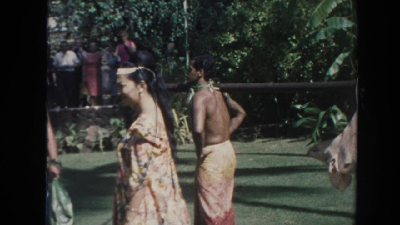 1961 Hawaii frame with a woman in a floral garment, a shirtless man, background onlookers, and a pig hanging from a pole.