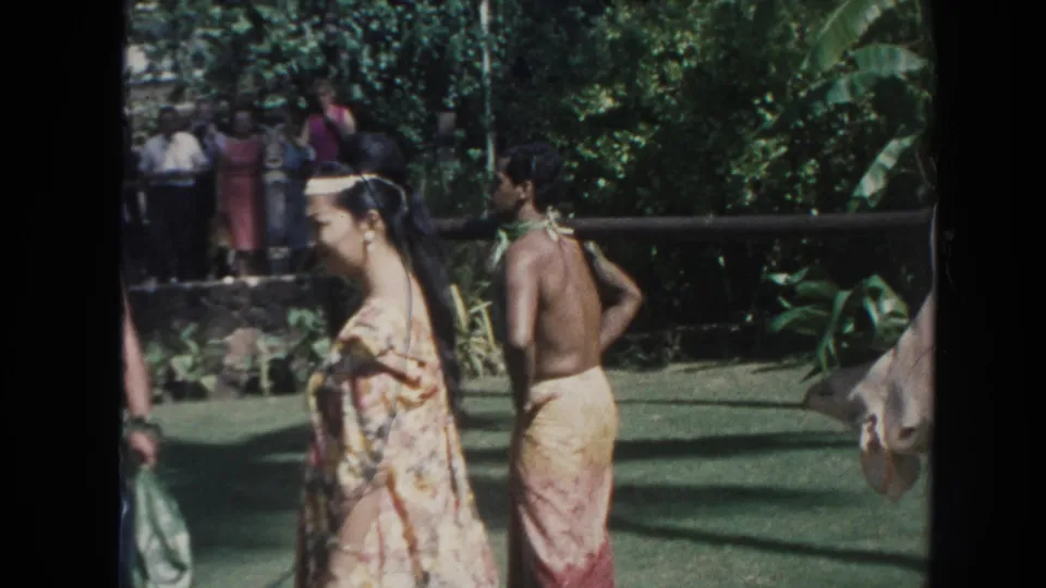 1961 Hawaii frame with a woman in a floral garment, a shirtless man, background onlookers, and a pig hanging from a pole.