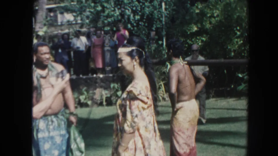 1961 Hawaii home-movie frame showing several adults near the preparation area, with tropical plants and onlookers behind them.