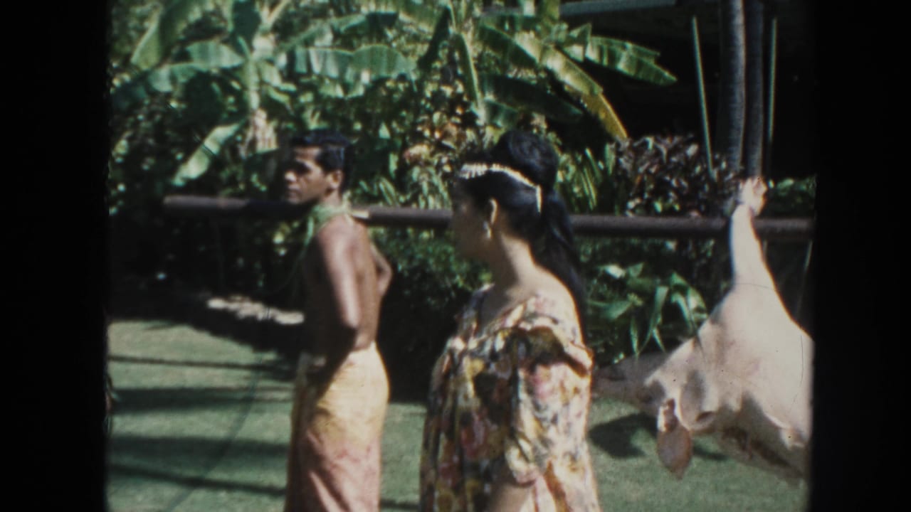 1961 Hawaii 8mm frame showing men in loincloth-style garments, a woman in a floral dress, and a white hog tied to a pole amid tropical foliage.