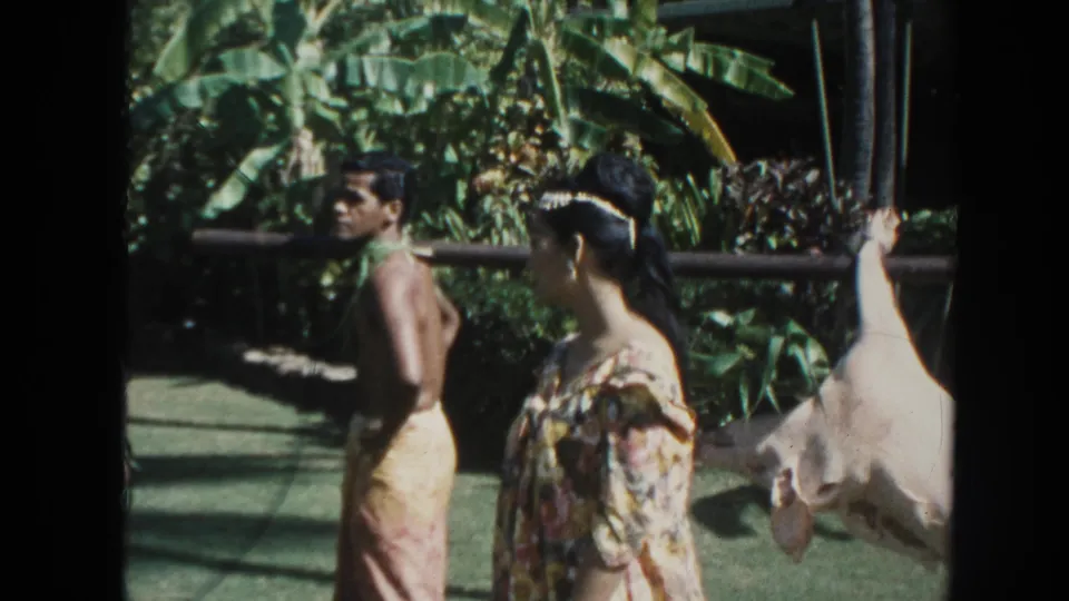 1961 Hawaii 8mm frame showing men in loincloth-style garments, a woman in a floral dress, and a white hog tied to a pole amid tropical foliage.