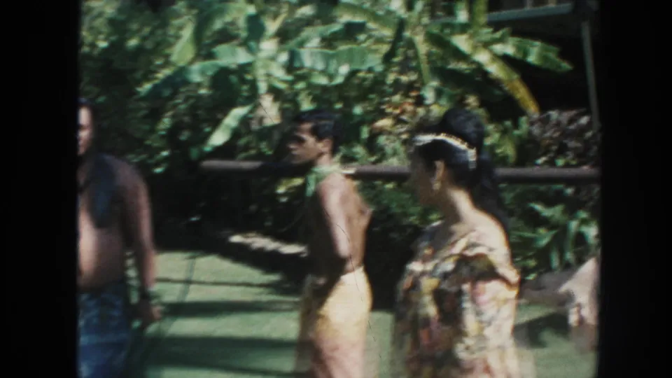 1961 Hawaii home-movie frame with men in traditional-style garments, a woman in floral clothing, and the tied hog partly visible at the edge.