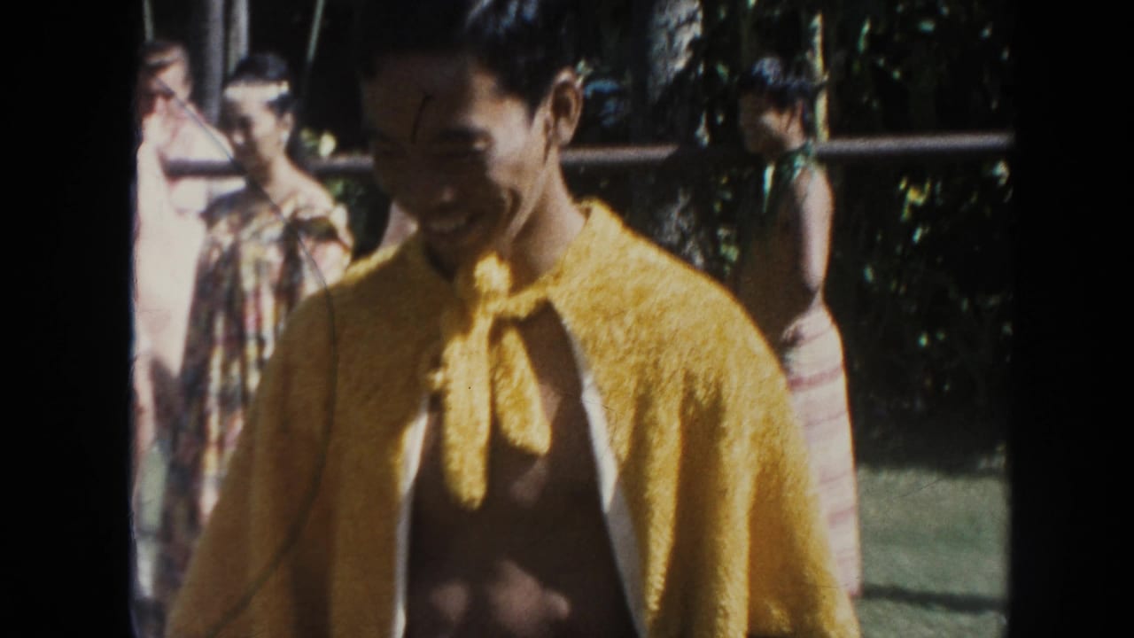 Smiling bare-chested man in a yellow cape stands outdoors with spectators behind him.